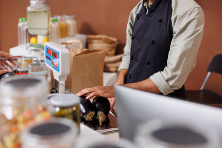 Selective focus on shopkeeper measuring two freshly harvested eggplants on modern weigh scale. Male vendor checking the weight and price of organic vegetables at cashier desk.の写真素材