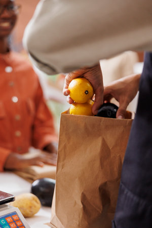 Close-up of male vendor placing freshly harvested produce into paper bag at checkout counter for female customer. Images shows person packing locally grown fruits and vegetables for takeaway.の写真素材