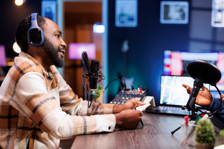 African american male journalist using his notebook and audio equipment to interview a person for his online talk show. Young black guy with a headset and speaking into a mic, is producing a podcast.の写真素材