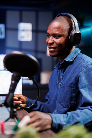 Black influencer speaking into microphone while creating online talk show at home is shown in...