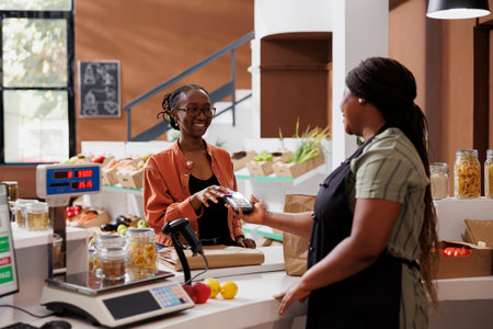 African American vendor serves a customer at a checkout counter using a debit card for wireless payment at eco friendly local store. Joyful client making cashless transactions at cashier desk.の写真素材