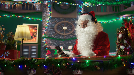 Cheerful Saint Nicholas listening to festive carols Christmas music on mobile phone, white bearded man spreading the winter holiday spirit in his log cozy cabin. Digital technology. Camera B.の写真素材