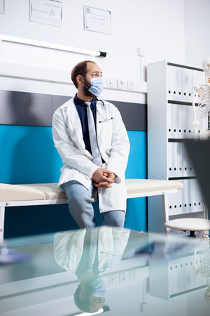 Young masked doctor with hands folded is alone in medical office waiting for patient appointments. Portrait of male general practitioner wearing lab coat and face mask, sitting on hospital bed.の写真素材