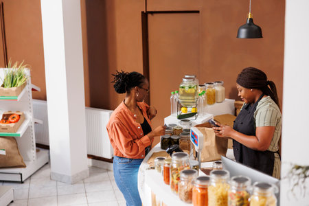 Black woman returning card into pocket after paying for bio food products and items she chose at an environmentally friendly store. Vendor and customer completing cashless transaction at store counterの写真素材