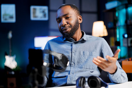 Audio engineer recording video for internet channel in dimly lit home studio, presenting pairs of gaming headphones to audience. Online star showing viewers music listening deviceの写真素材