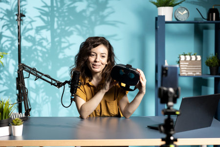 Smiling tech vlogger reviewing VR goggles in living room studio. Happy woman filming virtual reality...