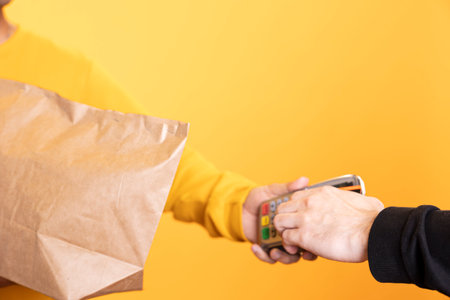 Client paying with credit card to part time delivery worker holding lunch meal order. Close up shot of man purchasing order in paper bag from courier, swiping card on POS device, studio backgroundの写真素材