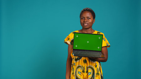 Optimistic gorgeous girl holding a pc with isolated green screen, posing with a smile in traditional ethnic clothes. Young relaxed woman presenting a mockup copy space display. Camera B.の写真素材