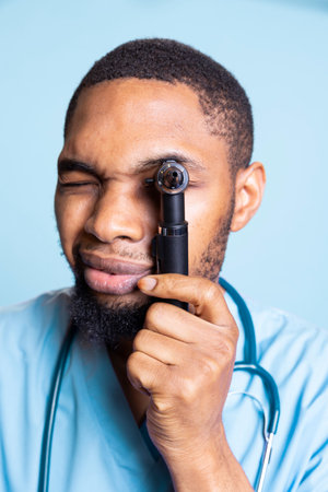 Nurse specialist does an ear drum examination to check the ear canal, doing otolaryngology checkup against blue background. African american male expert posing in studio.の写真素材