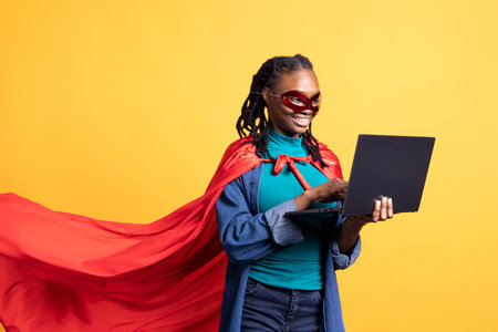 Smiling african american girl portraying superhero hiding identity, working on laptop, studio background. Jolly woman posing as hero in costume, solving tasks on notebook, covering face with maskの写真素材