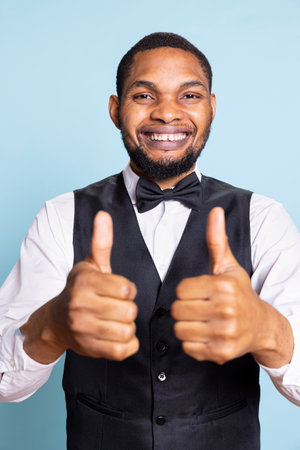 Smiling bellboy in suit and tie showing thumbs up gesture in studio, offering his approval and being satisfied. Positive doorkeeper gives a like sign, trust and professionalism.の写真素材