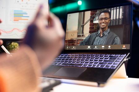 African american man talking on video call using personal computer, discussing about accounting infographics. Young adult communicating online and examining monthly revenue balance.の写真素材