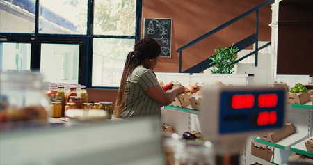 Loyal customer checking bio bulk products in zero waste shop, shopping for fresh natural groceries to restock pantry supplies. African american woman looks for vegan food. Handheld shot.の写真素材