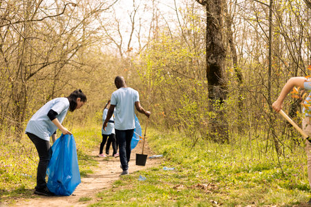 Diverse group of environmentalists collecting rubbish in trash bags, picking in junk and plastic waste from the forest. Activists cleaning the woods from garbage, nature pollution.の写真素材