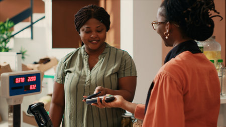 African american client buying eco produce with nfc payment, looking for homegrown pesticides free farming products at local farmers market. Woman paying with phone at pos terminal.の写真素材