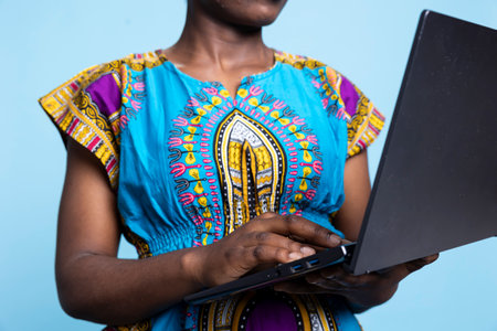 Young woman dressed in classic african american attire holds laptop in studio, surfing internet to explore webpages and social media platforms. Smiling, modern woman enjoys wifi technologies.の写真素材