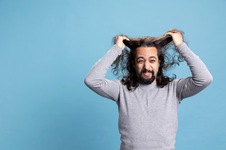 Middle eastern guy acting desperate and pulling his hair out on camera, being furious and stressed against blue background. Young adult feeling overwhelmed and in despair, distress.の写真素材