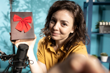 Joyful content creator holding present box, excited to do giveaway for subscribers, filming with selfie camera. POV shot of media star happy to offer packed gift with ribbon to viewersの写真素材