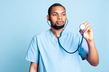 African american specialist listening to heartbeat and deep breaths at consultation with a patient, cardiology exam against blue background. Young nurse in scrubs with stethoscope.の写真素材