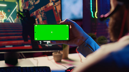 Man in dimly lit apartment watching tutorial on how to finish videogame on green screen smartphone, enjoying day off. Gamer learning online multiplayer shooter tips from video on mockup mobile phoneの写真素材