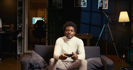 Gamer arriving home, ready to start game and enjoy leisure time. African american man sitting on couch in living room, playing videogame on gaming console, relaxing after workの写真素材