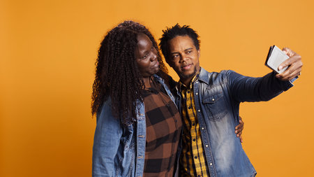 African american husband and wife having fun taking selfies together on phone against yellow background, enjoying leisure time. Joyful confident married people laughing for pictures. Camera B.の写真素材