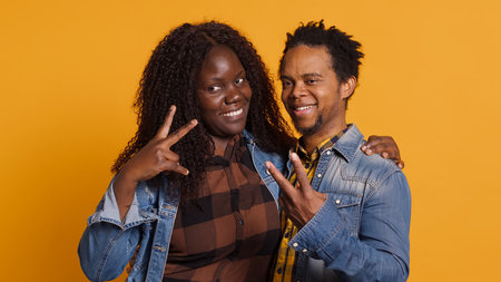 Cute life partners posing in studio and doing peace sign in studio, showing the victory symbol against yellow background. African american spouses doing kawaii gesture. Camera A.の写真素材