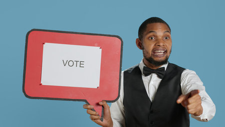 Bellboy hotelier showing a speech bubble with the message to vote. Hotel porter staff in tuxedo holds a cardboard icon.の写真素材