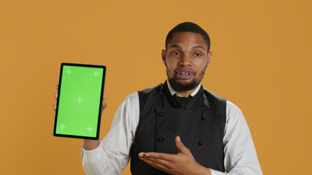 Restaurant waiter presenting a green screen layout on tablet against yellow background, wearing professional service oriented attire on duty. Butler serving clients, professionalism. Camera A.の写真素材