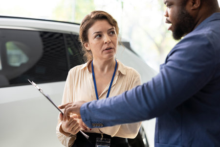 Dealership saleswoman answering man questions about cars, financing and purchase process. Vehicle showroom agent explaining african american client warranties, services and available extra featuresの写真素材