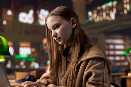 Intellectual college student researching public relations and its societal impact, utilizing her laptop and educational materials from the university library archives to complete a class assignment.の写真素材