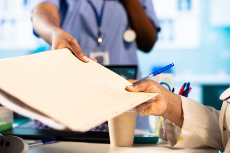 Female nurse brings the patient clinical records for a checkup routine with medic practitioner, offering advice and guidance for wellbeing. Black man at the medical cabinet. Close up.の写真素材