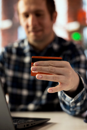 Focus on credit card used by man in blurry background paying bills, confirming payments on laptop. Close up of debit card used by person at home filling taxes online using notebookの写真素材
