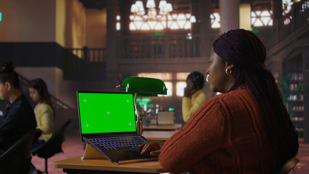Black librarian typing on laptop with chroma key in a quiet public library, dedicated to organizing...