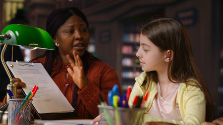 African american professor mentoring diverse schoolgirls in the library, learning together in a...