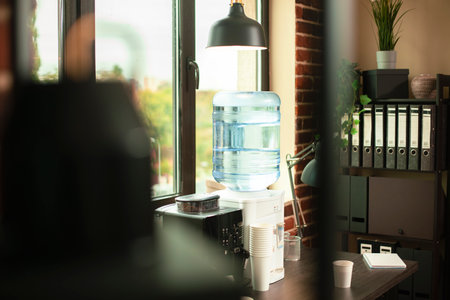 Corner office space having coffee machine, water cooler, and plastic cups positioned beside folder cabinet. Refreshments and file binders neatly arranged in brightly lit brick wall room.の写真素材