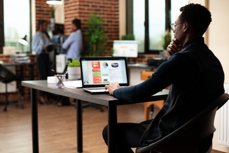African American man analyzing company performance and market data using laptop in office. Male professional focusing on financial reports and conducting a research on his device in modern workspace.の写真素材