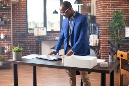 African american manager placing laptop on desk in brick wall office, ready to begin work for the day. Businessman standing by table and positioning his personal computer at startup company workspace.の写真素材
