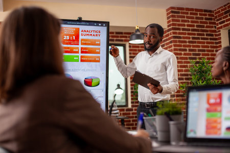 Businessman with clipboard in hand, using digital monitor to explain analytics summary in meeting. African american entrepreneur doing project presentation, planning marketing strategy with his team.の写真素材