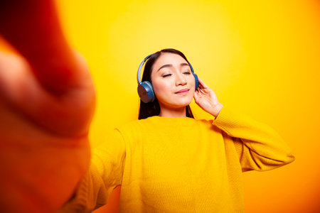 Young woman listening to music with her eyes closed and making a daily vlog for her viewers. Asian lady wearing headphones and holding camera for a selfie against isolated yellow background.の写真素材