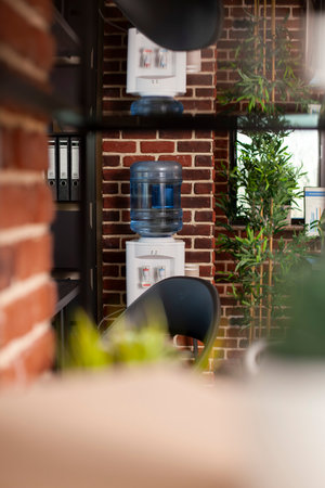 Selective focus of water cooler set near office cabinet and plant decoration by brick wall. Modern workspace arranged with indoor greenery and refreshment area for comfortable and productive workday.の写真素材