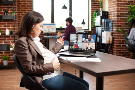 Professional expectant mother with hand on belly bump, conducting online meeting with team on laptop. Pregnant woman sitting at desk with notebook and speaking to coworkers on videocall on device.の写真素材