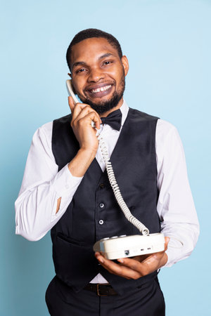 Professional doorkeeper staff makes a room reservation on landline phone call, discussing about hotel amenities and facilities to help guests. Bellboy with suit and tie answers remote call.の写真素材