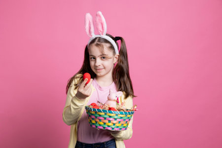Cheerful lovely kid showing colorful easter decorations in a basket, holding painted eggs and a pink stuffed rabbit. Young sweet girl posing with bunny ears and festive cute ornaments.の写真素材