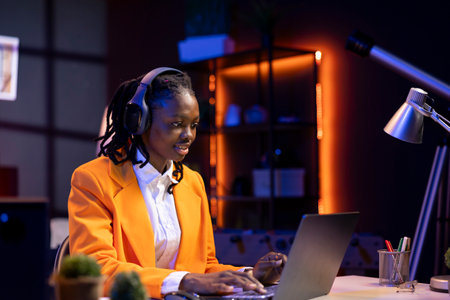 Remote worker listening music, analyzing data on laptop at desk, compiling analytical reports. Woman in home office wearing headphones, using notebook device, typing informationの写真素材