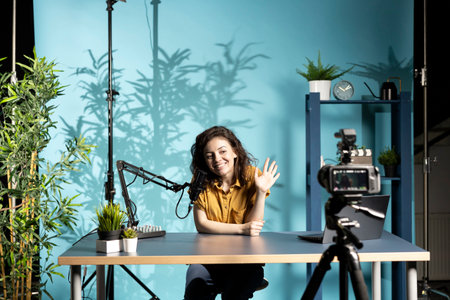 Jolly woman greeting fans, filming vlog with high tech gear in professional studio, speaking into microphone equipment. Smiling influencer saluting subscribers, talking about trending topics on online channelの写真素材