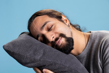 Sleepy young man taking a nap with a pillow standing against blue background, pretending to fall...