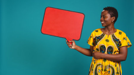 Woman in ethnic clothes holds a speech bubble carton board to create an ad commercial, presenting an isolated billboard icon in studio. Young adult posing for advertisement. Camera B.の写真素材
