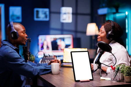 On desk, white screen with mockup template is displayed on tablet while African American vloggers are recording live podcast while wearing headsets. Digital device on table shows isolated chromakey.の写真素材