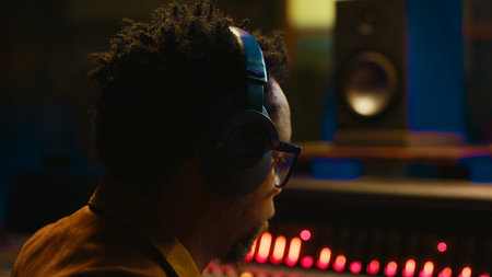 African american sound engineer using headphones to listen to audio recordings in post production, adjusting volume levels with editing software interface. Expert in studio control room. Camera A.の写真素材